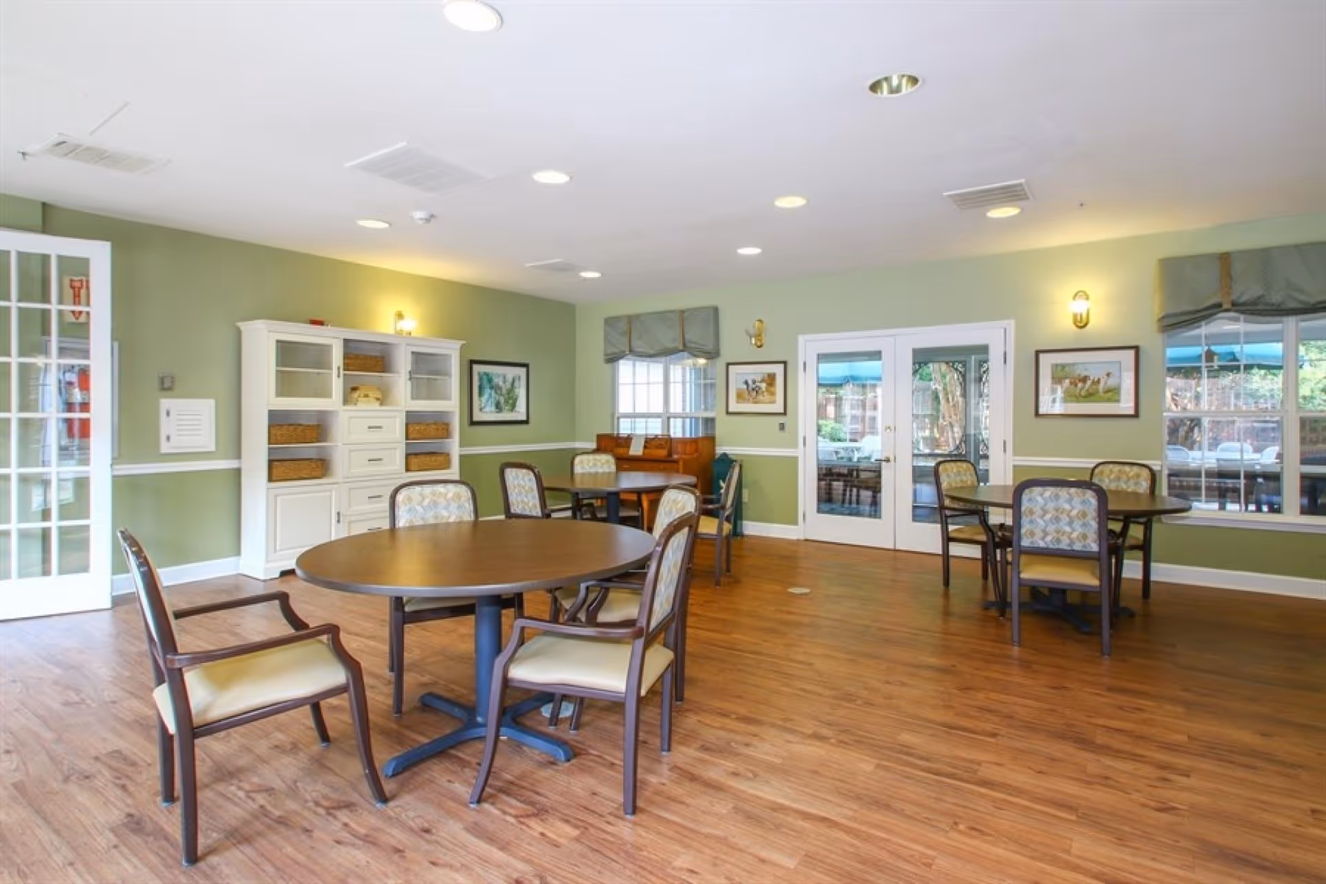 Well-lit communal dining room with round tables and chairs, wood floors, green walls, and glass doors and windows.