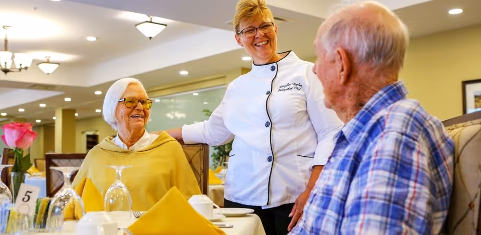 A smiling staff member in a chef jacket chats with two elderly residents seated at a set dining table in a communal dining room.
