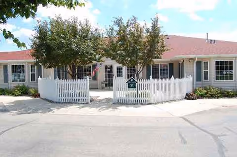 Front view of a single story memory care facility with a white picket fence and trees.