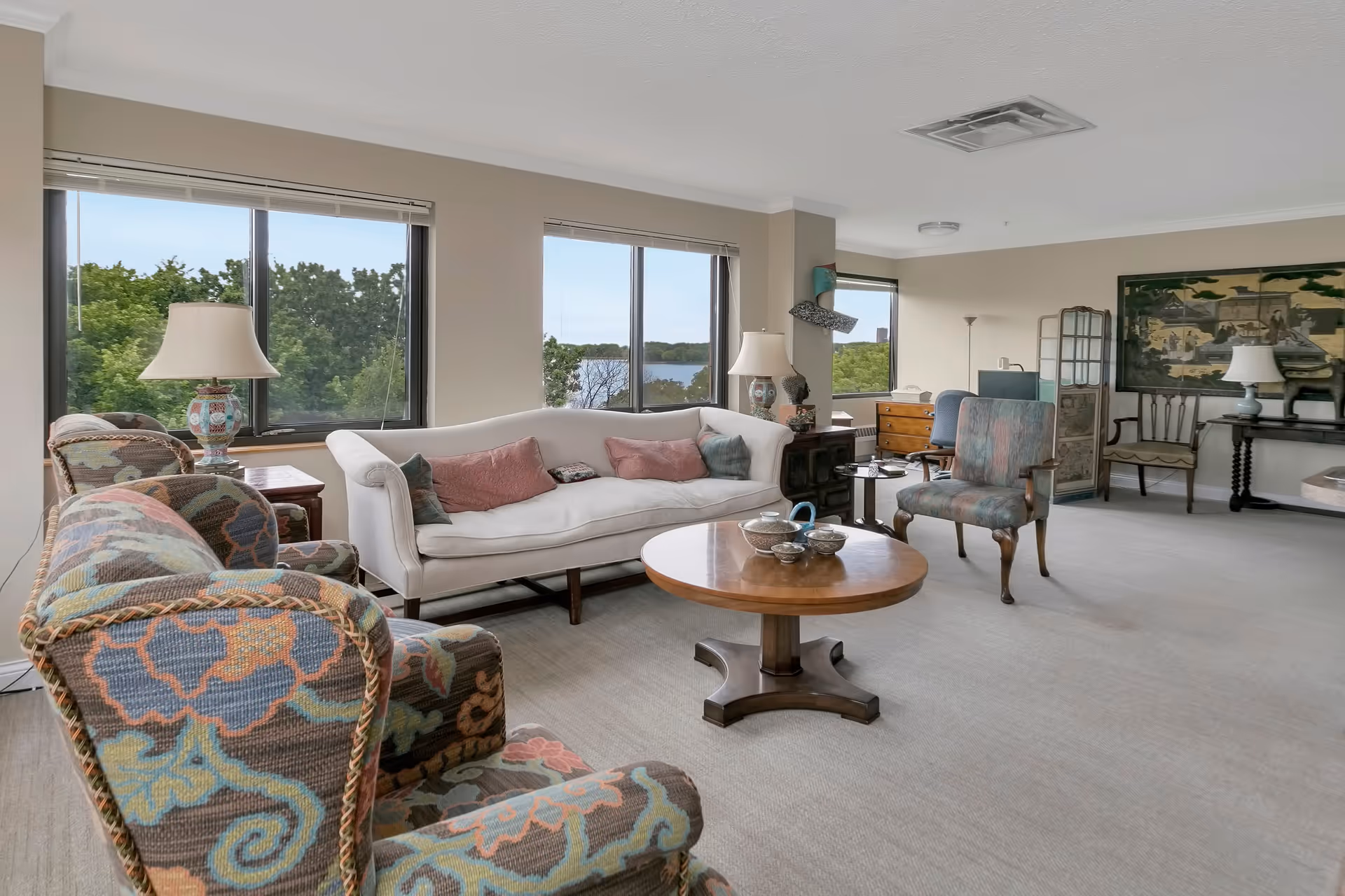 A spacious senior living room with large windows showing a view of trees and a lake. The room features a white sofa with pink and green cushions, two patterned armchairs, a round wooden coffee table with tea set, a wooden side table with a decorative lamp, a desk with a chair, and various decorative items including a large framed artwork on the wall.