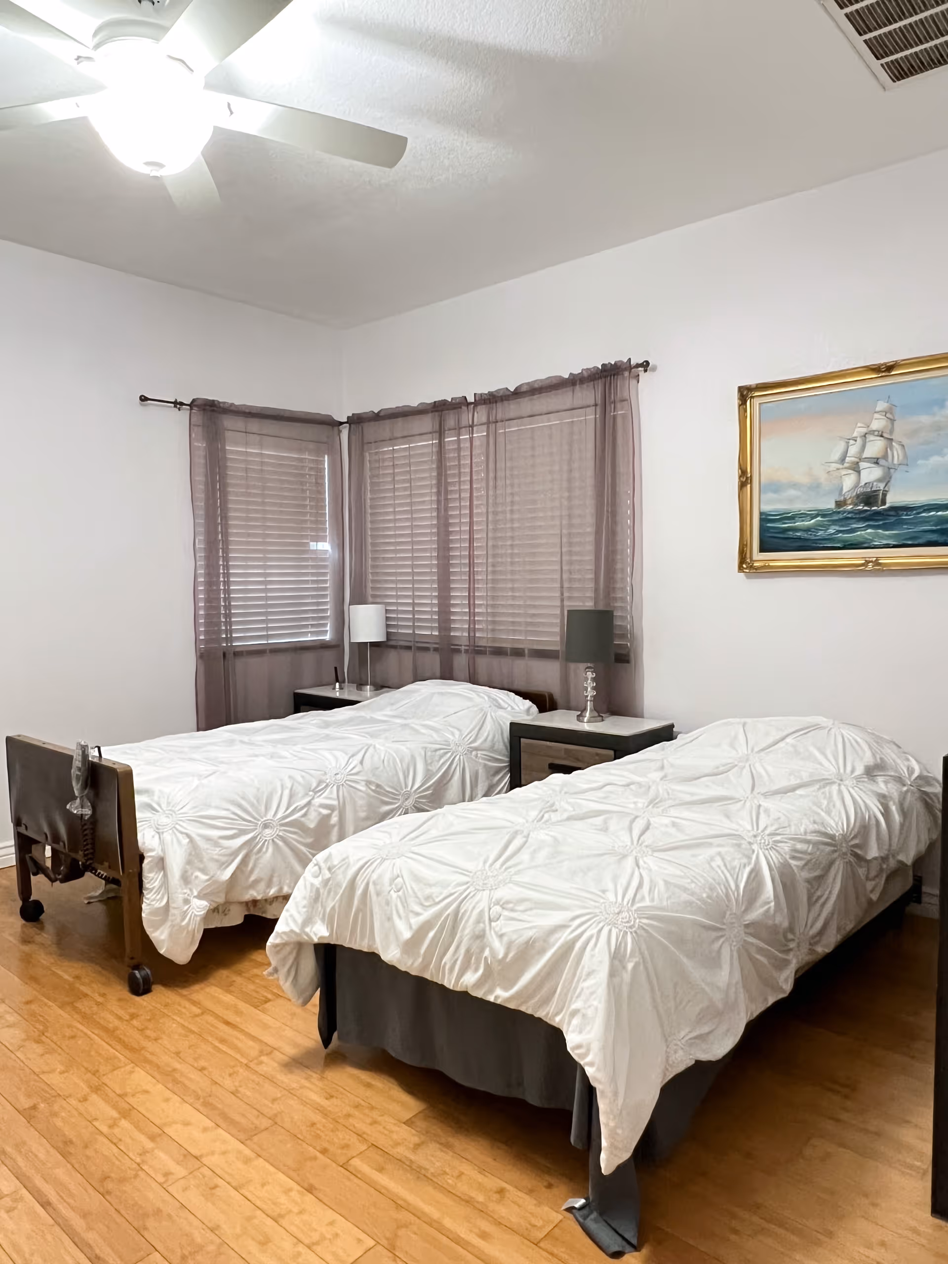 A bright bedroom with two twin beds, nightstands and lamps, a ceiling fan, and a framed ship painting on the wall.