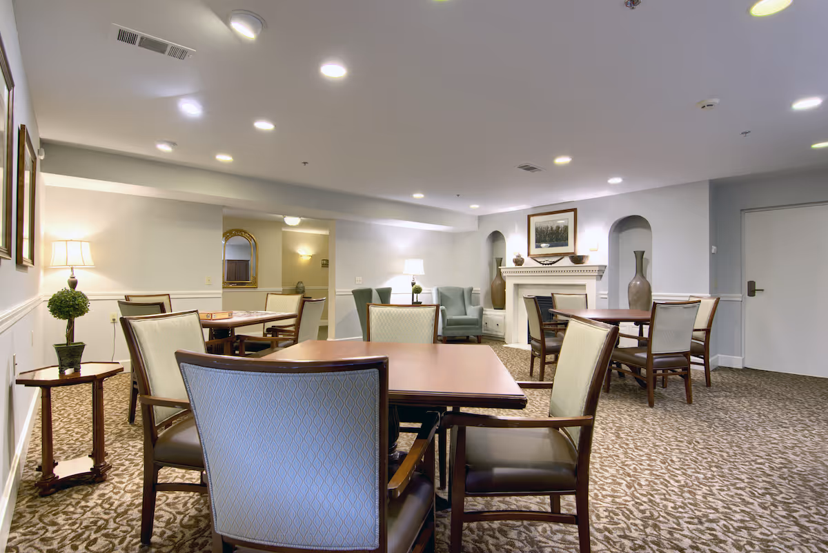 Well-lit senior living common room with multiple dining tables and upholstered chairs, a seating area and decorative fireplace.