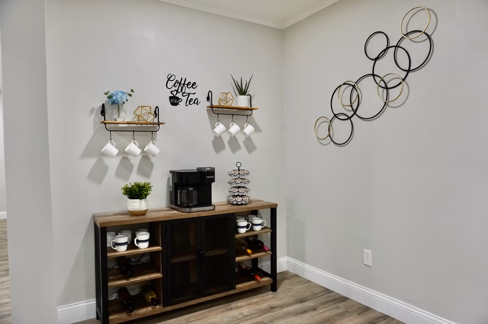 A coffee and tea station in a senior living facility with a coffee maker on a wooden cabinet, shelves holding white mugs, decorative plants, and a wall sign that reads 'Coffee and Tea'. The wall is light gray with a modern circular metal wall art decoration.