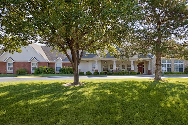 Front exterior of a single-story brick and siding senior living building with a covered entrance and a grassy, tree-shaded lawn in front.