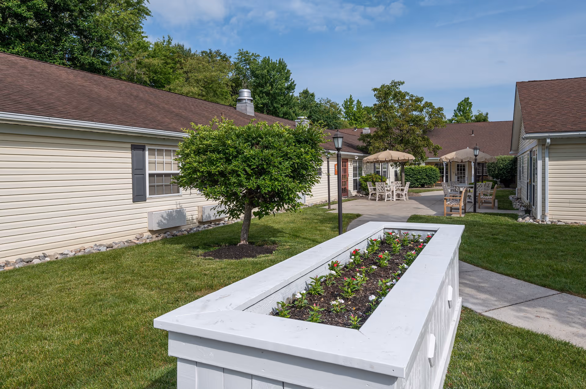 Outdoor courtyard area at Arden Courts - ProMedica Memory Care Community with green grass, a small tree, a white rectangular planter box with flowers, paved walkways, and patio tables with umbrellas surrounded by beige buildings with brown roofs.