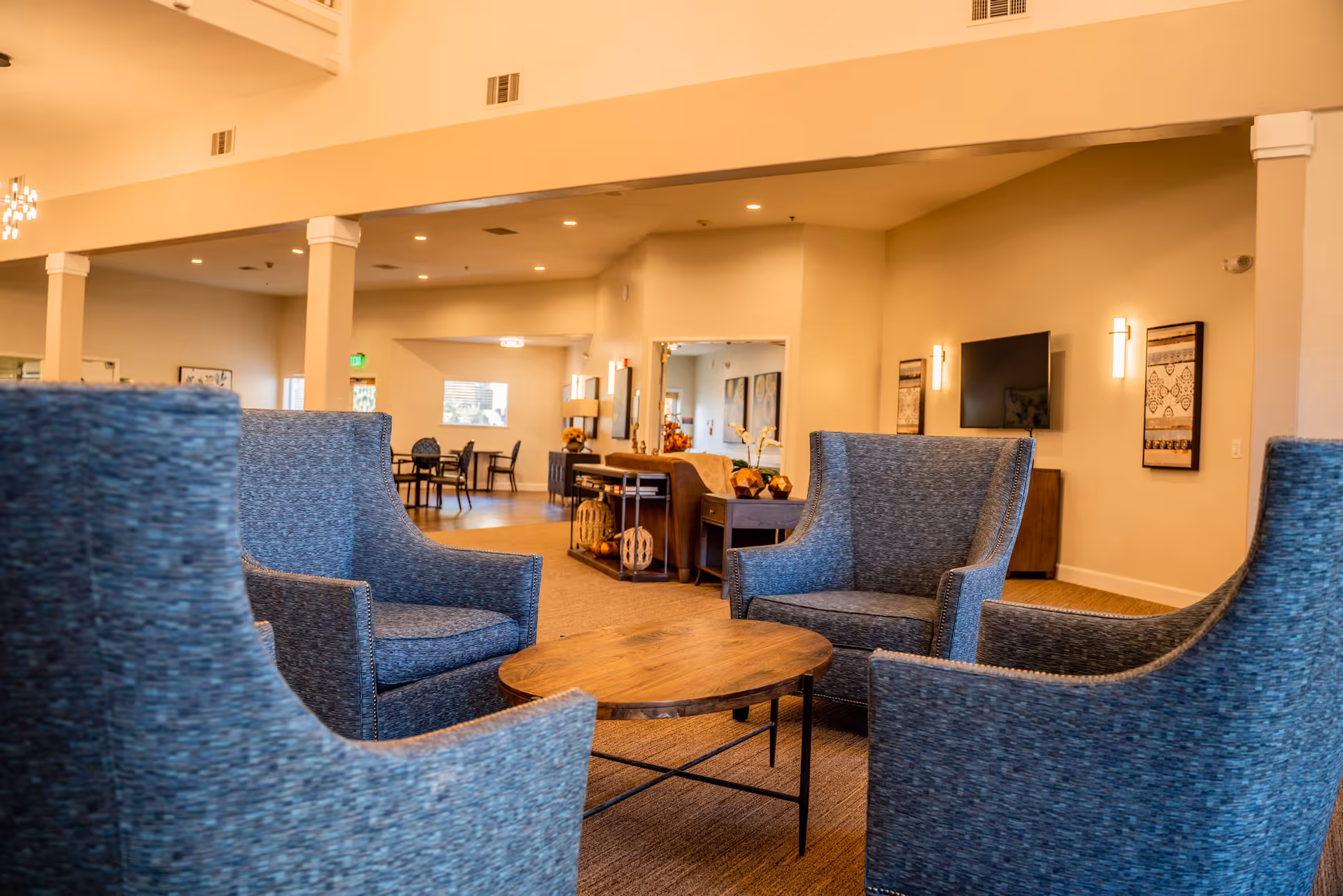 A cozy seating area in a senior living facility with four blue upholstered armchairs arranged around a round wooden coffee table. The room has beige walls, carpeted floors, and warm lighting. In the background, there is a dining area with tables and chairs, wall-mounted TV, and decorative artwork on the walls.