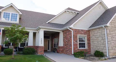 Exterior view of a single-story building with a combination of brick, stone, and siding walls. The building has a covered entrance supported by columns, a small tree and bushes in front, and a concrete walkway leading to the door. The address number 2895 is visible on one of the columns.