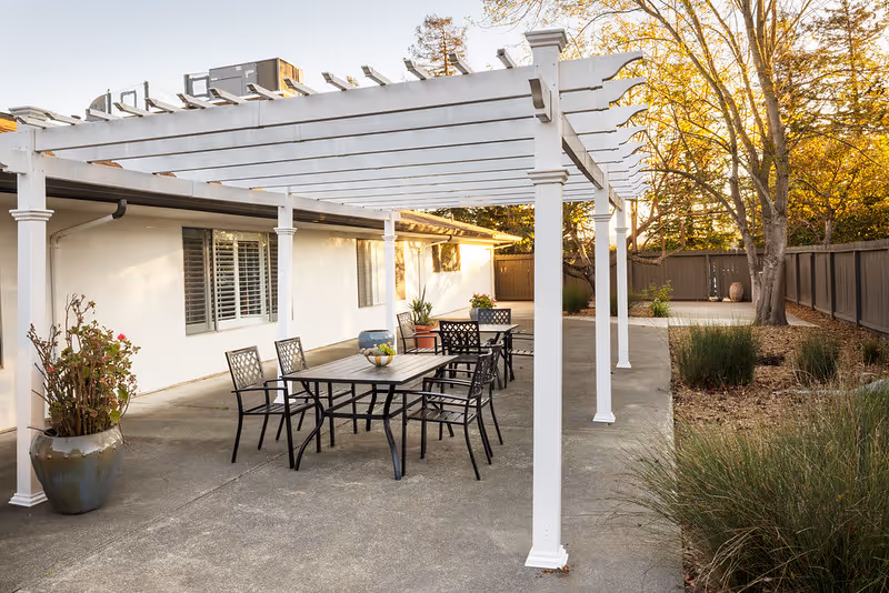 Outdoor patio area with a white pergola covering two black metal tables and several matching chairs. There are potted plants near the building wall and along the edge of the patio. The area is surrounded by a wooden fence and trees with autumn foliage.