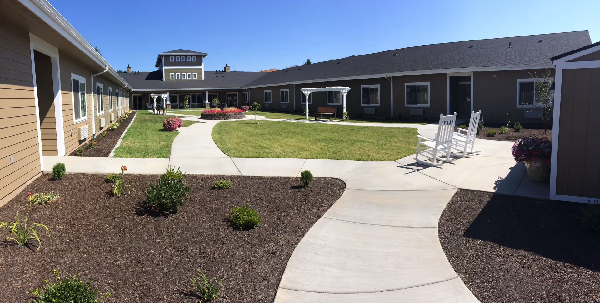 Outdoor courtyard area of Fieldstone Memory Care Yakima with a paved walkway, green lawn, small garden beds with plants, two white rocking chairs, a bench under a white pergola, and a building surrounding the courtyard under a clear blue sky.