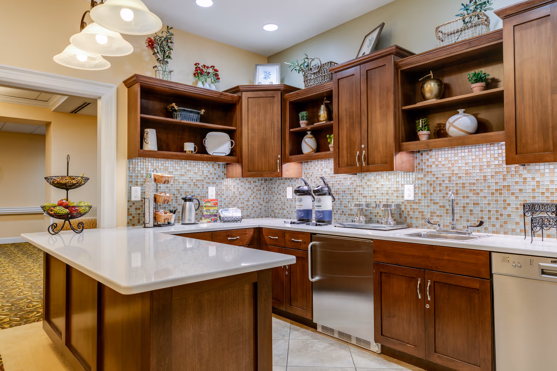 A well-lit kitchen area with wooden cabinets, a white countertop island, mosaic tile backsplash, and stainless steel appliances including a dishwasher. The shelves hold decorative items and plants, and there are two beverage dispensers on the counter. A fruit basket is visible on the left side near the entrance to another room.