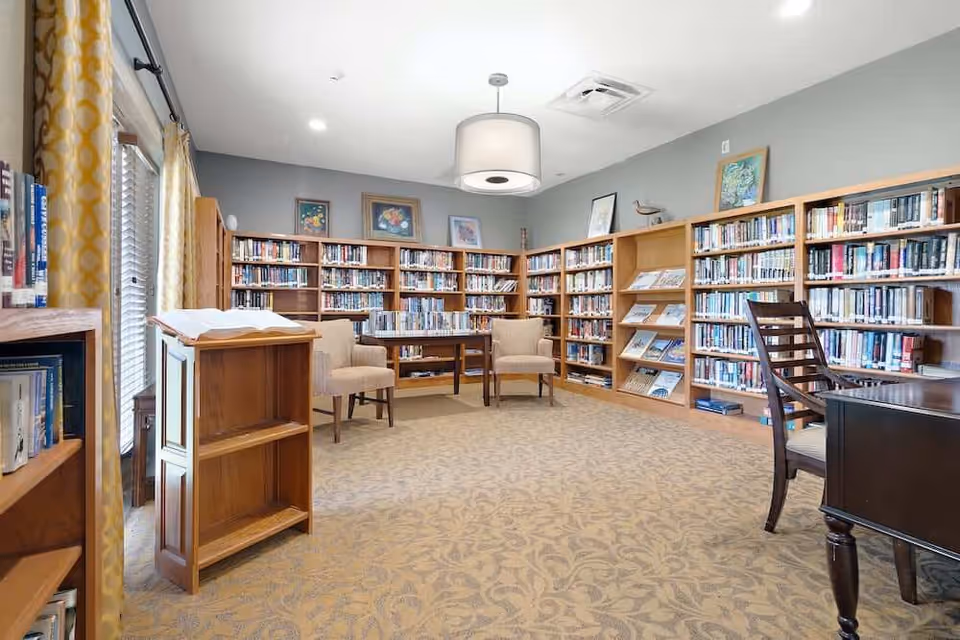 A well-lit library room with bookshelves filled with books along the walls, two beige armchairs around a small wooden table, a wooden lectern with an open book, a wooden desk with a chair, and framed artwork on the walls.