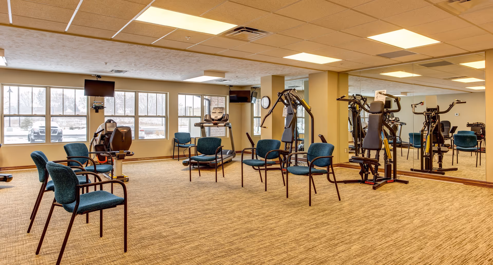 A fitness room in a senior living facility with exercise equipment including a recumbent bike, treadmill, and weight machines. Several teal upholstered chairs are arranged in a circle in the center of the room. Large windows let in natural light, and a wall of mirrors reflects the room. Two wall-mounted televisions are visible.