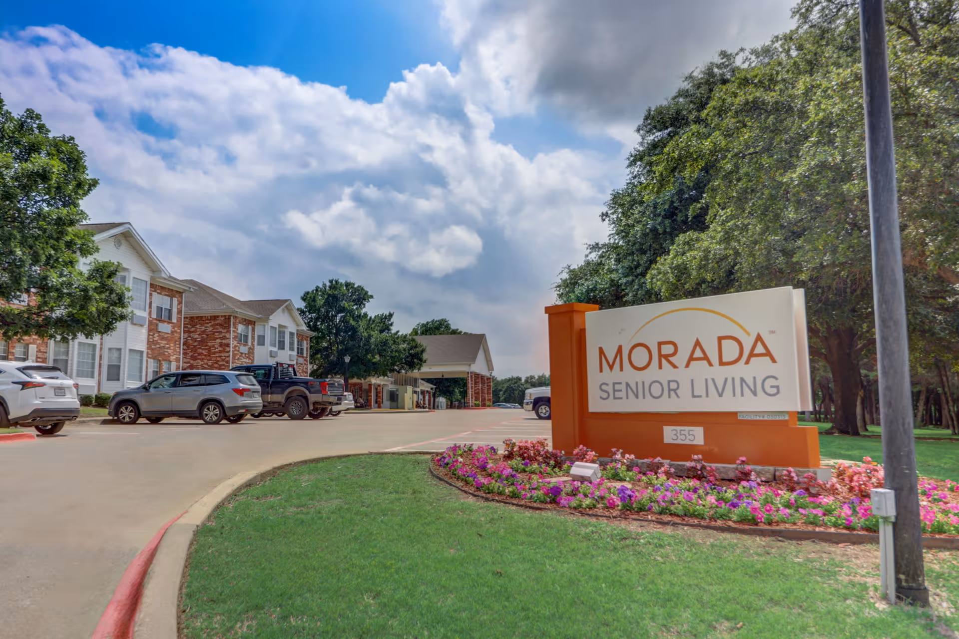 Entrance to Morada Senior Living facility with a large sign displaying the name. The sign is surrounded by colorful flowers and green grass. In the background, there are parked cars and a two-story brick and white building under a partly cloudy sky.