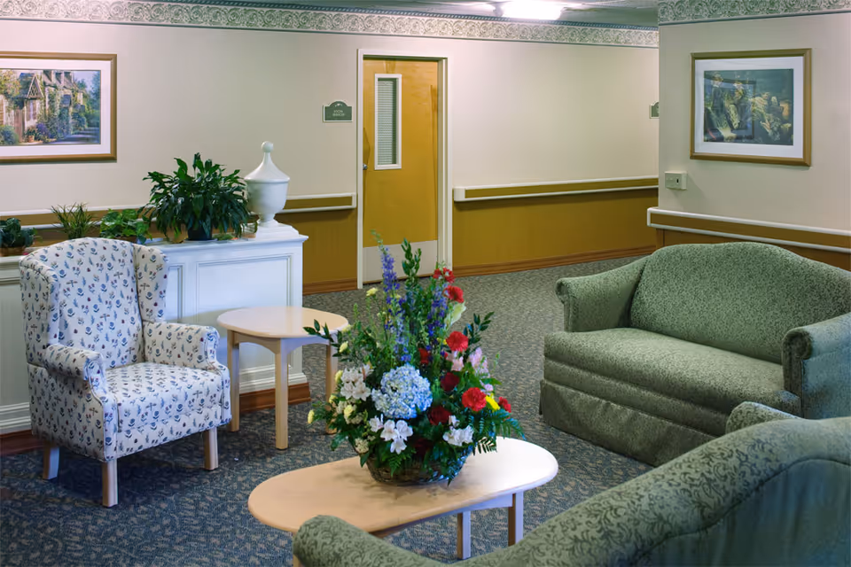 A cozy sitting area in a senior living facility with two green upholstered sofas, a floral patterned armchair, and two light wooden tables. A large floral arrangement is placed on the central table. The walls are decorated with framed paintings and there are several potted plants on a white cabinet. A door labeled 'Social Services' is visible in the background.