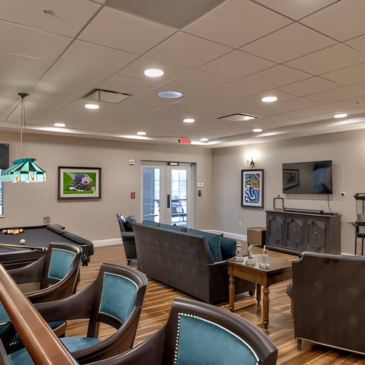 A spacious and well-lit living room area in an assisted living facility featuring comfortable seating including armchairs and sofas arranged around a wooden coffee table. A flat-screen TV is mounted on the wall above a decorative cabinet. The room has wooden flooring, framed artwork on the walls, and a pool table with a green hanging lamp above it. Double glass doors lead to an outdoor area.