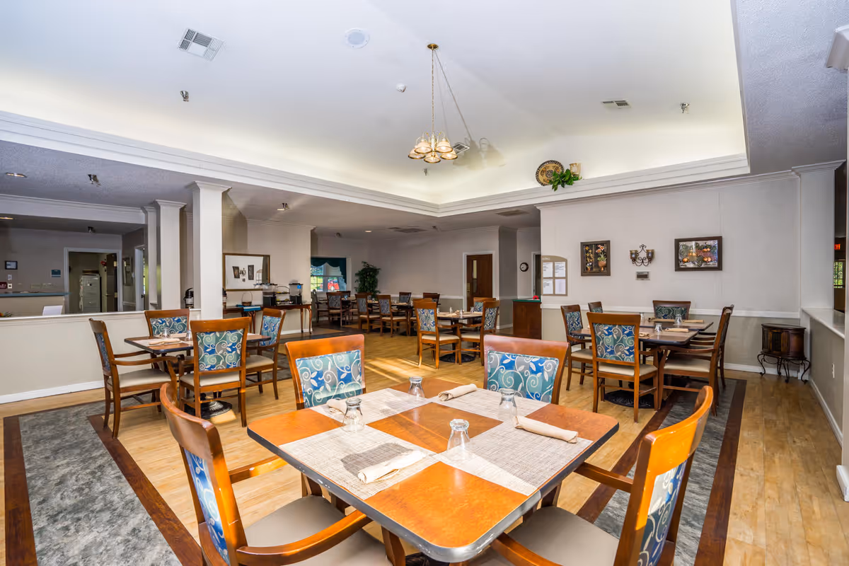 A dining room in Saunders House featuring multiple wooden tables with patterned cushioned chairs. Each table is set with placemats, napkins, and upside-down glasses. The room has a light wood floor, neutral-colored walls, and a ceiling with recessed lighting and a central chandelier. There are framed pictures and a small plant on the walls, and a sideboard with additional chairs and items in the background.