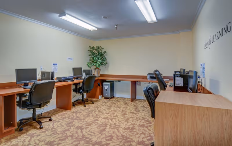 A computer room with several desktop computers on wooden desks arranged along the walls. There are black office chairs at each workstation, a potted plant in the corner, and a patterned carpet on the floor. The walls are light yellow, and fluorescent lights illuminate the room. A partial wall decal reads 'Learning'.