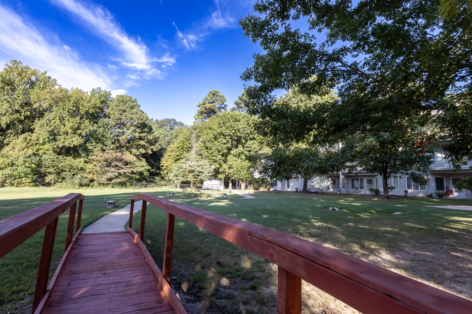 Red wooden footbridge leading to a grassy courtyard with large trees and a multi-unit building under a blue sky.
