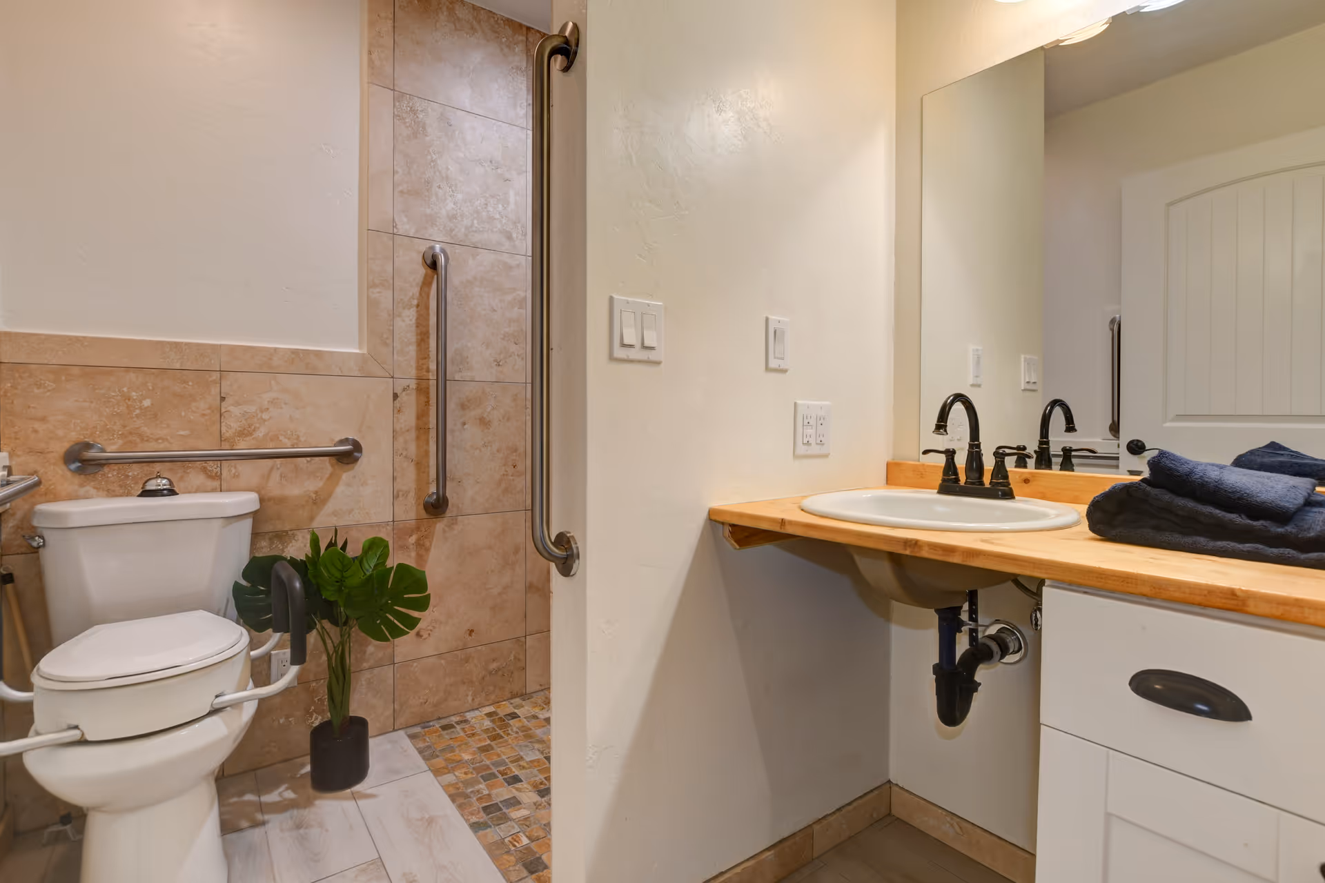 Accessible bathroom with a white toilet equipped with safety rails, a small potted plant beside it, a walk-in shower with tiled walls and grab bars, and a wooden countertop with a sink and folded dark towels next to a large mirror.