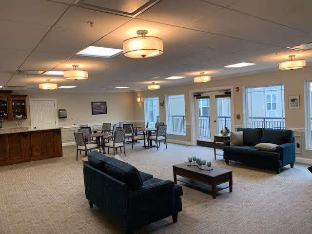 A spacious common area with two dark blue sofas facing each other with a wooden coffee table in between. Behind the sofas are several tables and chairs arranged for seating. The room has large windows and glass doors letting in natural light, beige walls, and ceiling lights with round fixtures. There is a wooden cabinet on the left side of the room.