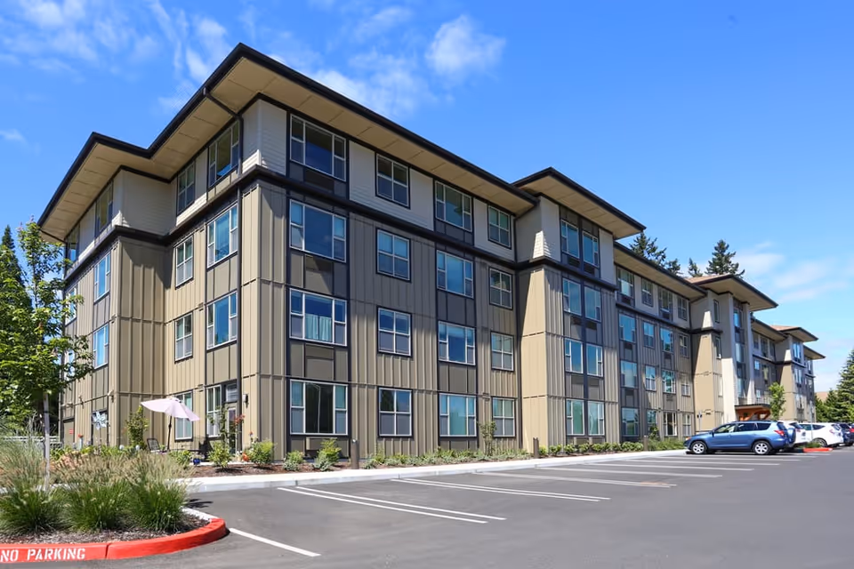 Exterior view of a modern four-story senior living facility building with multiple windows, a parking lot with several cars, and landscaping including small trees and bushes under a clear blue sky.