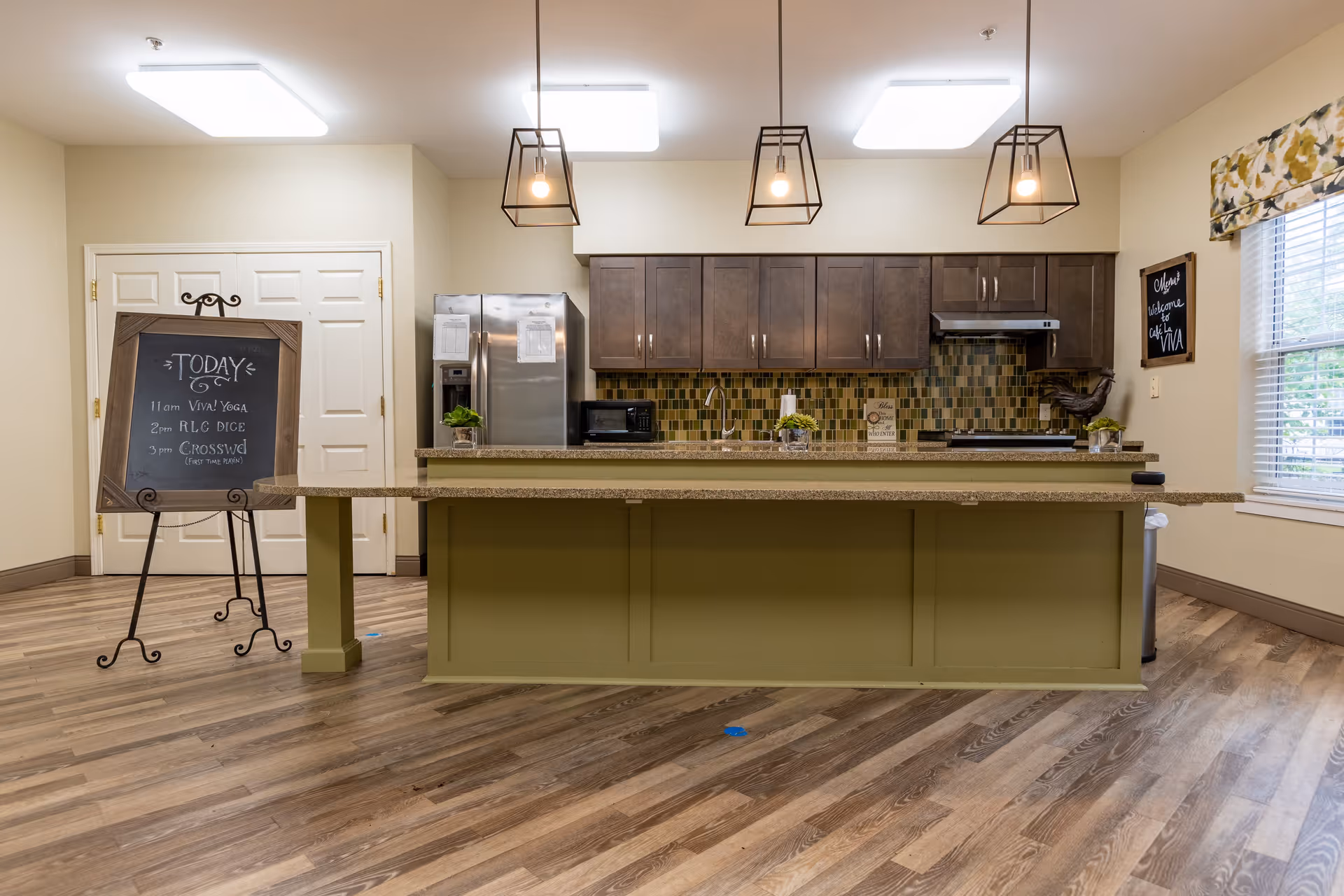 A modern kitchen area in Walnut Ridge Assisted Living and Memory Care featuring a large island with a green base and granite countertop, three pendant lights hanging above, dark wood cabinets, a stainless steel refrigerator, microwave, and stove. There is a chalkboard on an easel listing daily activities and a window with floral valance on the right side.
