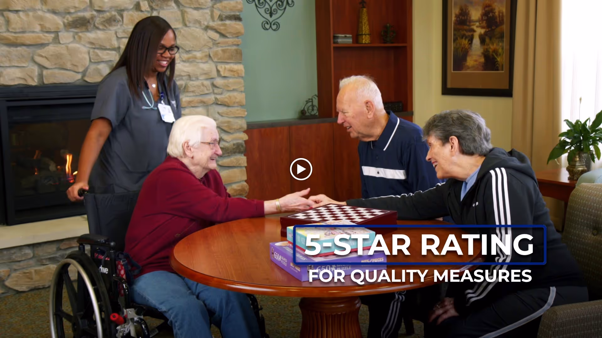 A nurse stands smiling next to an elderly woman in a wheelchair who is shaking hands with another elderly woman seated at a round wooden table. An elderly man is also seated at the table. They appear to be in a cozy room with a stone fireplace, wooden shelves, and a framed picture on the wall.