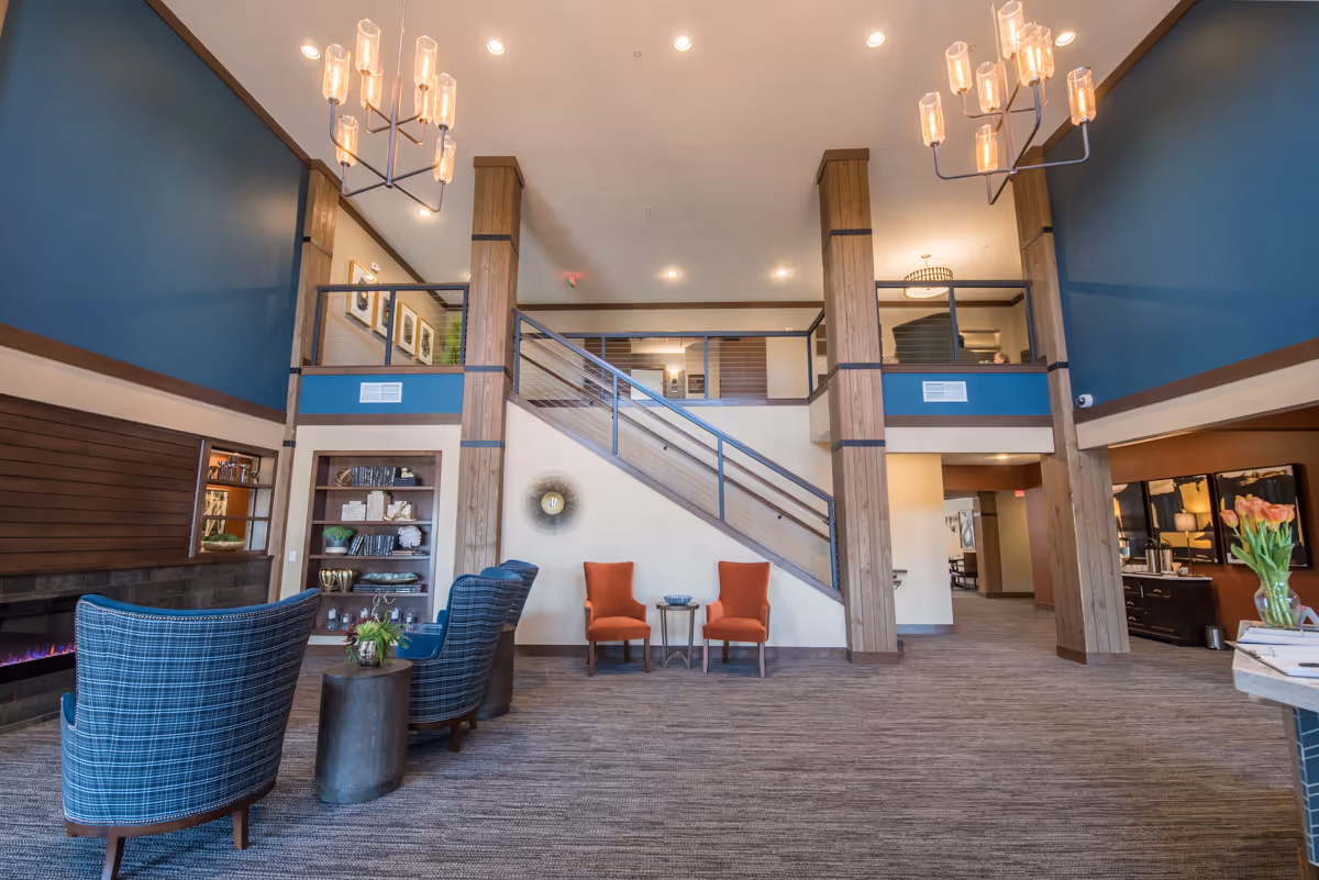 Spacious and well-lit common area with high ceilings featuring two modern chandeliers. The room has a staircase with metal railings leading to an upper level. There are two orange armchairs with a small table between them against the wall under the stairs. On the left side, there are two blue plaid armchairs with a small round table between them, positioned near a long modern fireplace. Wooden columns and blue accent walls add warmth and style to the space. Shelves with decorative items and framed pictures are visible in the background.
