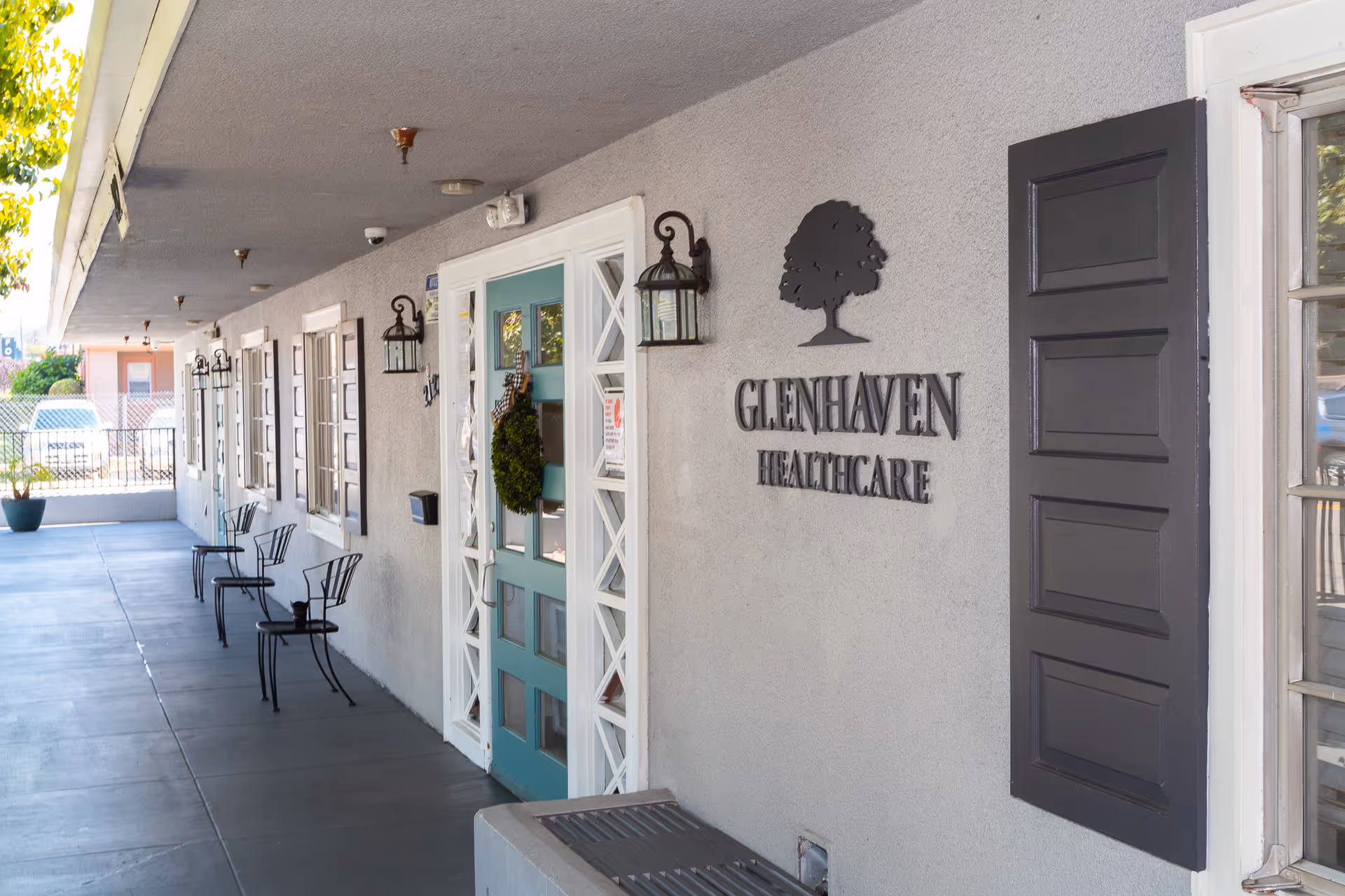 Exterior view of Glenhaven Healthcare facility showing a covered walkway with several black metal chairs and a teal door decorated with a wreath. The building has gray walls with black shutters and wall-mounted lantern-style lights. The Glenhaven Healthcare sign with a tree logo is mounted on the wall near the entrance.