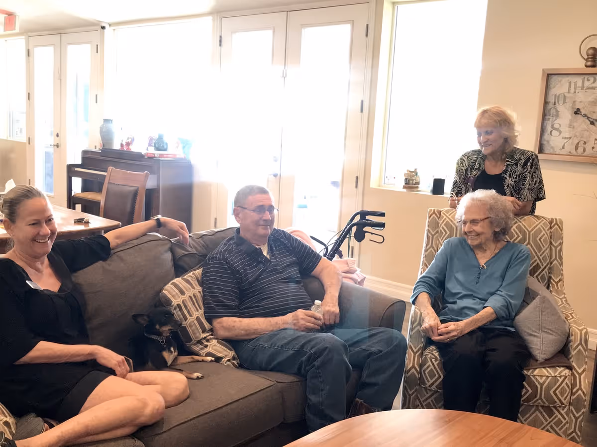 A group of four people sitting and standing in a bright living room area. Two elderly individuals are seated on patterned armchairs, a man is sitting on a gray couch holding a water bottle with a small dog beside him, and a woman is standing behind one of the armchairs. The room has large windows and a clock on the wall.