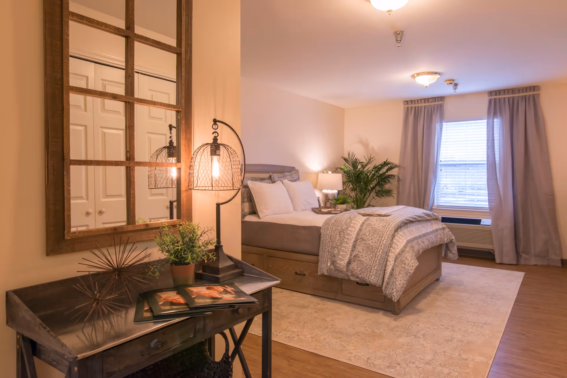 A cozy bedroom in a senior living facility featuring a neatly made bed with patterned bedding, a large window with curtains, a bedside table with a lamp and a plant, and a wooden console table with a decorative lamp, a potted plant, and photo albums. A large framed mirror hangs above the console table.
