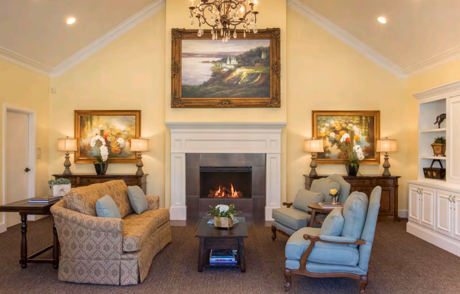A cozy living room with a lit fireplace centered on a cream-colored wall. Above the fireplace hangs a large framed painting of a scenic landscape. On either side of the fireplace are matching wooden side tables with lamps and floral paintings above them. The room features a patterned beige sofa with blue cushions on the left and two blue upholstered armchairs with wooden arms on the right. A dark wooden coffee table with decorative items and books is placed in the center on a carpeted floor. Built-in white cabinetry is visible on the right side of the room.