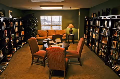 A cozy reading room with a mustard yellow sofa against the back wall under a window, a round wooden table with four upholstered chairs in the center, flanked by tall bookshelves filled with books on both sides, and a table lamp on a side table next to the sofa providing warm lighting.