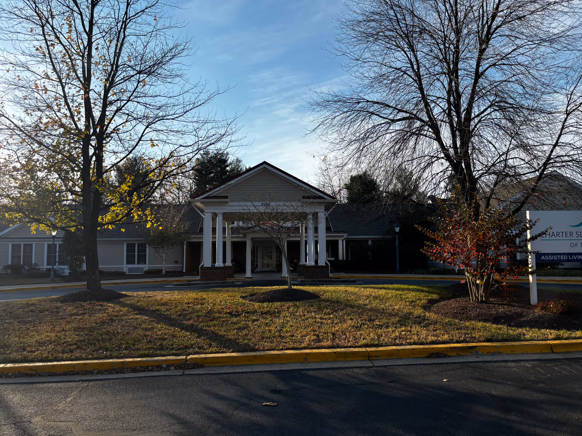 Front exterior view of Charter Senior Living Of Bowie building with a covered entrance supported by white columns, surrounded by trees with some autumn leaves and a lawn area in front.