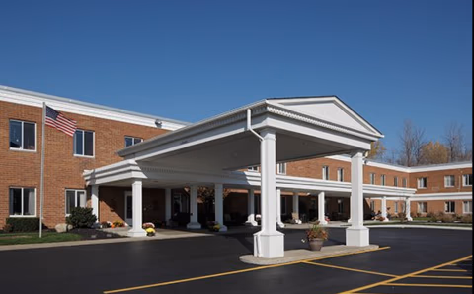 Exterior view of Heathwood Assisted Living and Memory Care building with a covered entrance supported by white columns, an American flag on a flagpole, and a clear blue sky.