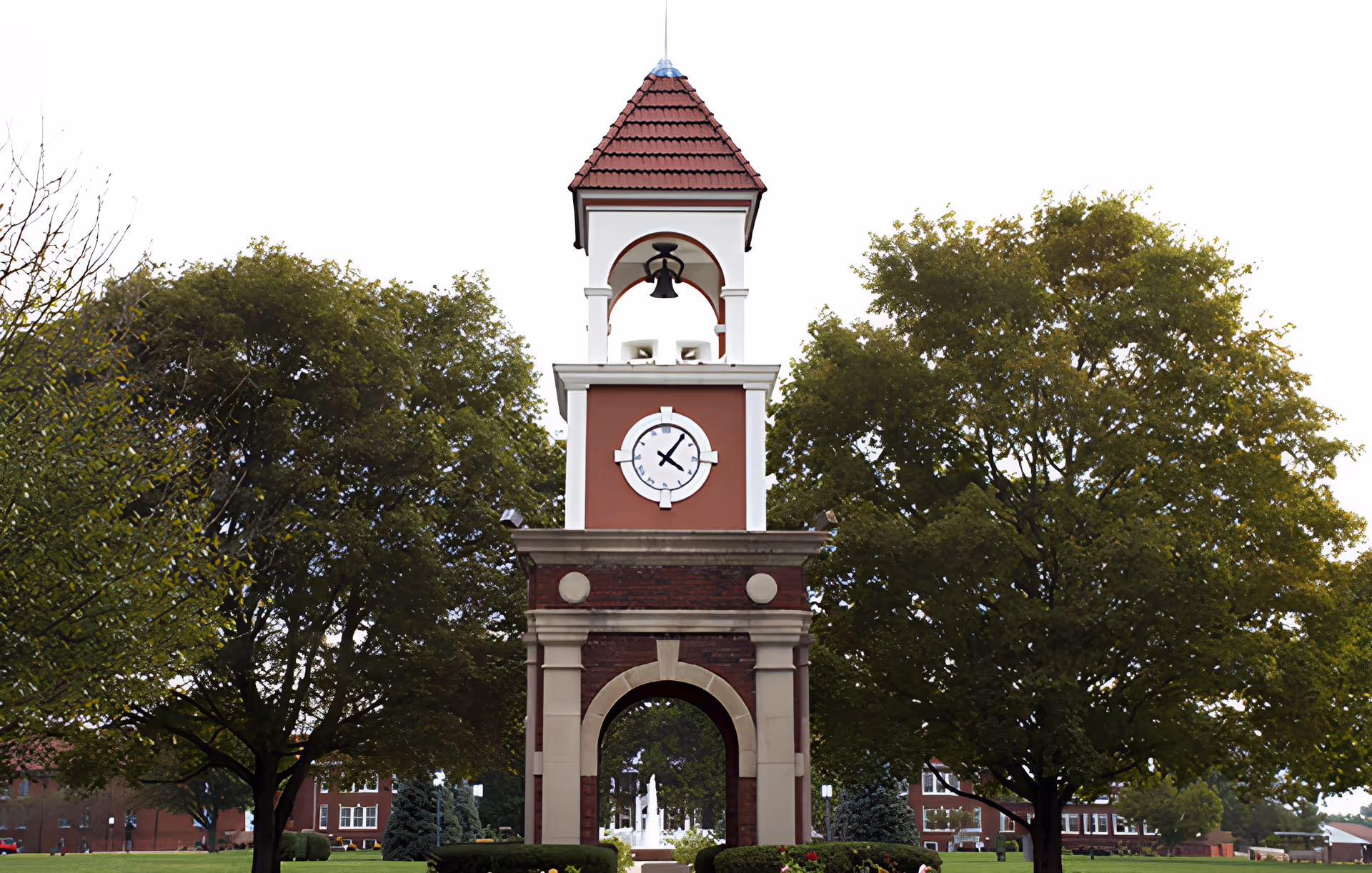 A tall clock tower with a red tiled roof and a bell under an arch, surrounded by green trees and a well-maintained lawn with buildings in the background.