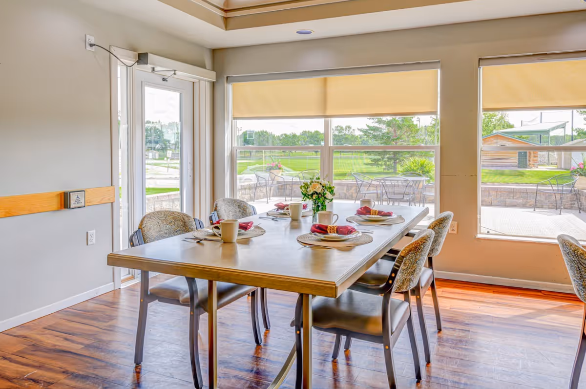A dining area with a rectangular table set for four people, featuring plates, cups, napkins, and a small flower arrangement in the center. The room has large windows with beige roller shades, allowing natural light to brighten the space. Outside the windows, there is a patio with metal chairs and tables, and a green lawn with trees and a wooden structure in the background.