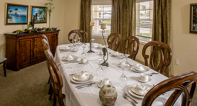 Formal dining room with a long table set for a meal, wooden chairs, a sideboard, and windows with curtains.