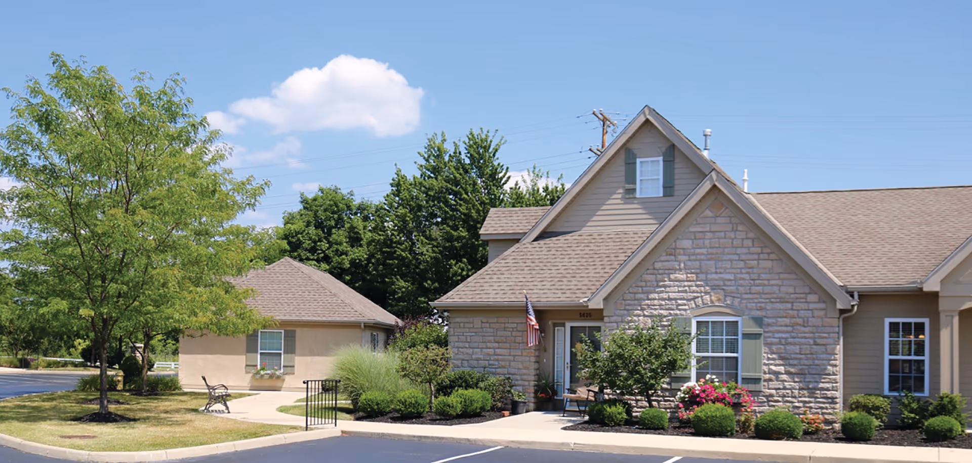 Exterior view of a senior living facility named Feridean Commons featuring a stone and siding building with a peaked roof, surrounded by landscaped bushes and trees under a blue sky with some clouds. There is a small paved parking area in front and a bench near the entrance.