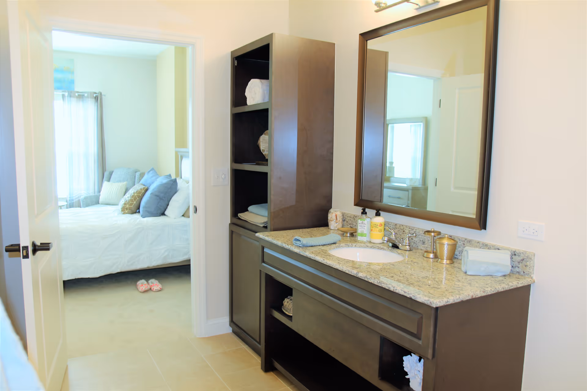 A bathroom vanity with a granite countertop, a sink, a large mirror, and a dark wood cabinet with shelves holding towels and decorative items. In the background, an open door reveals a bedroom with a bed, multiple pillows, a window with curtains, and a pair of slippers on the floor.