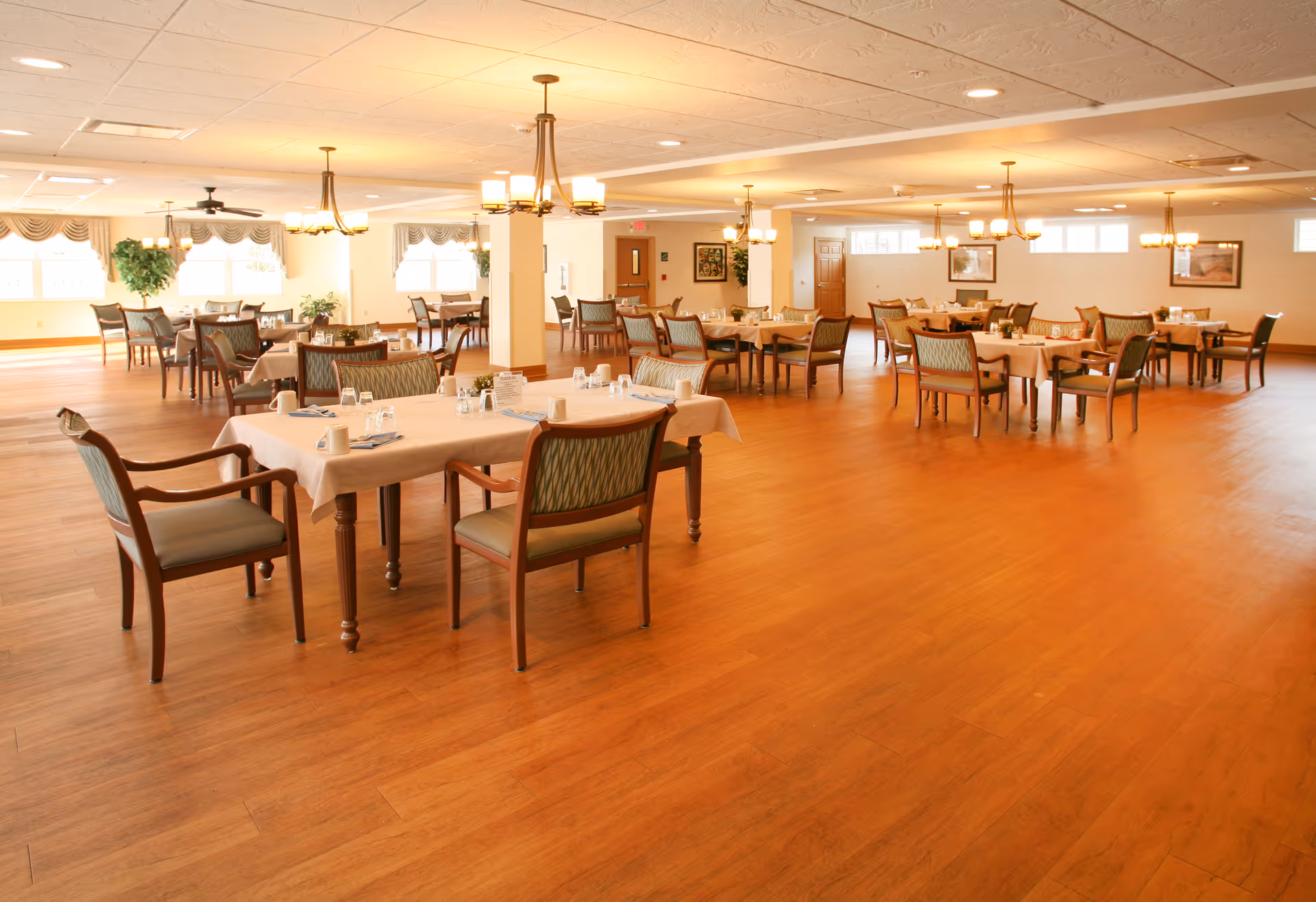 Spacious dining room with multiple set tables and chairs on a wood floor and overhead chandeliers.