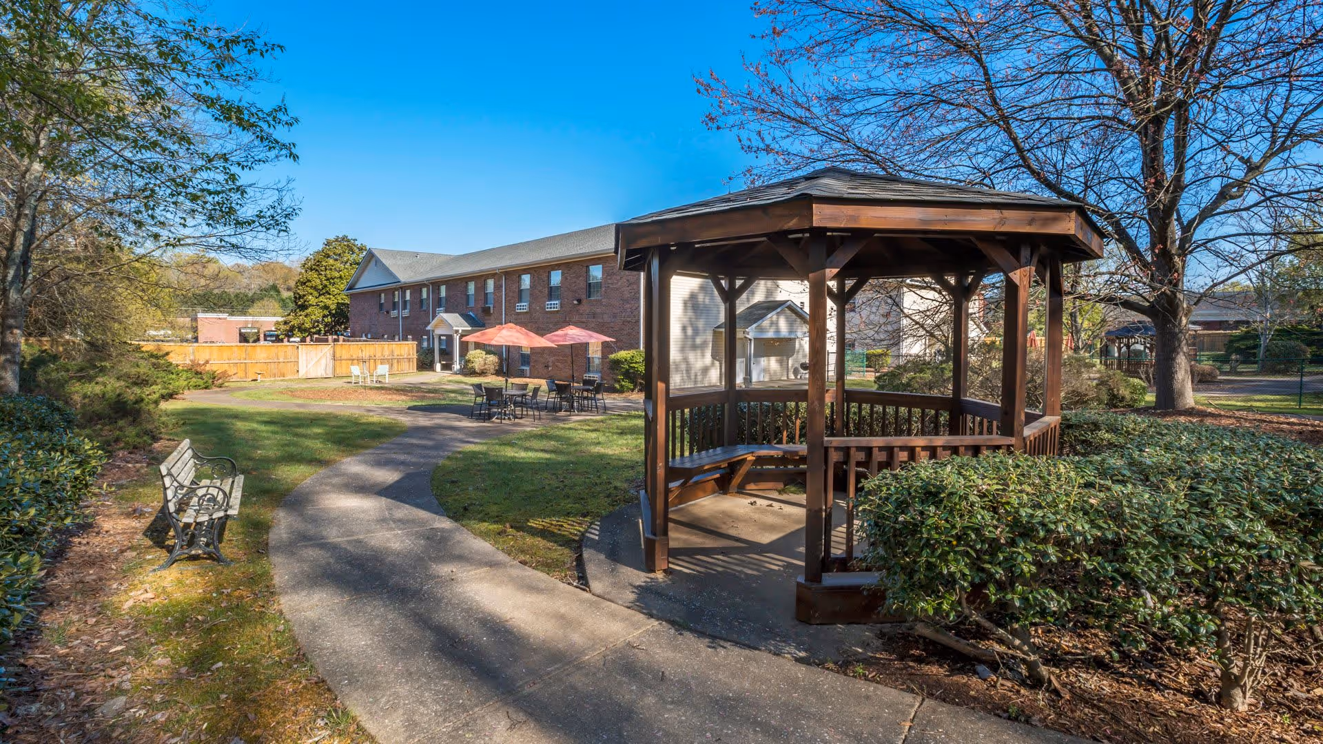 Outdoor garden area at American House Hamilton Place featuring a wooden gazebo with built-in benches, a curved concrete pathway, a metal bench, patio tables with umbrellas, and surrounding trees and bushes under a clear blue sky.