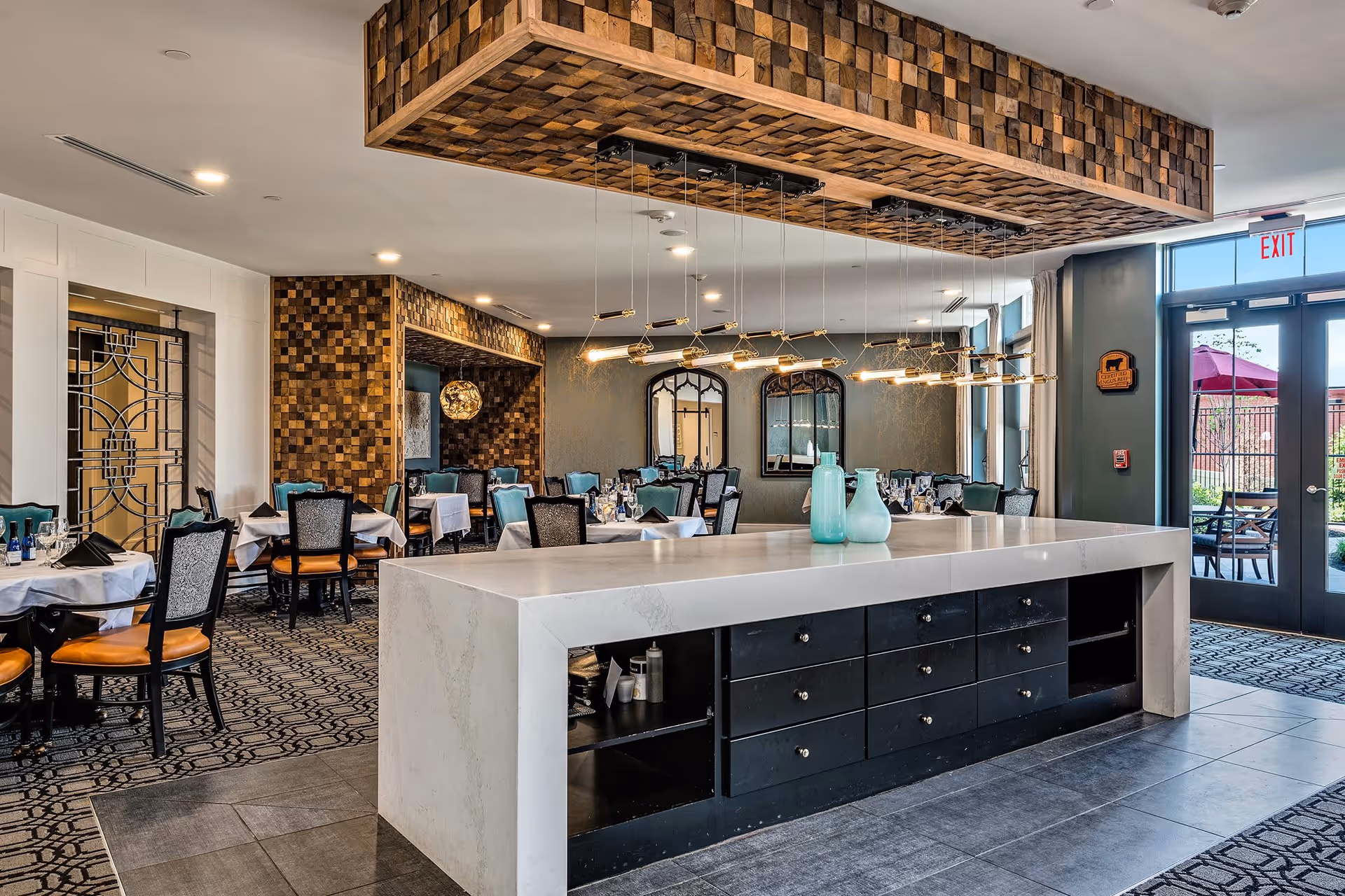 A spacious and elegant dining room with multiple tables covered in white tablecloths and set with glassware and napkins. The room features a large central island with a white countertop and black drawers, decorated with two blue vases. Above the island hangs a modern light fixture with multiple bulbs. The walls have decorative wood paneling and mirrors, and large windows and glass doors provide natural light and a view of an outdoor patio area with red umbrellas.