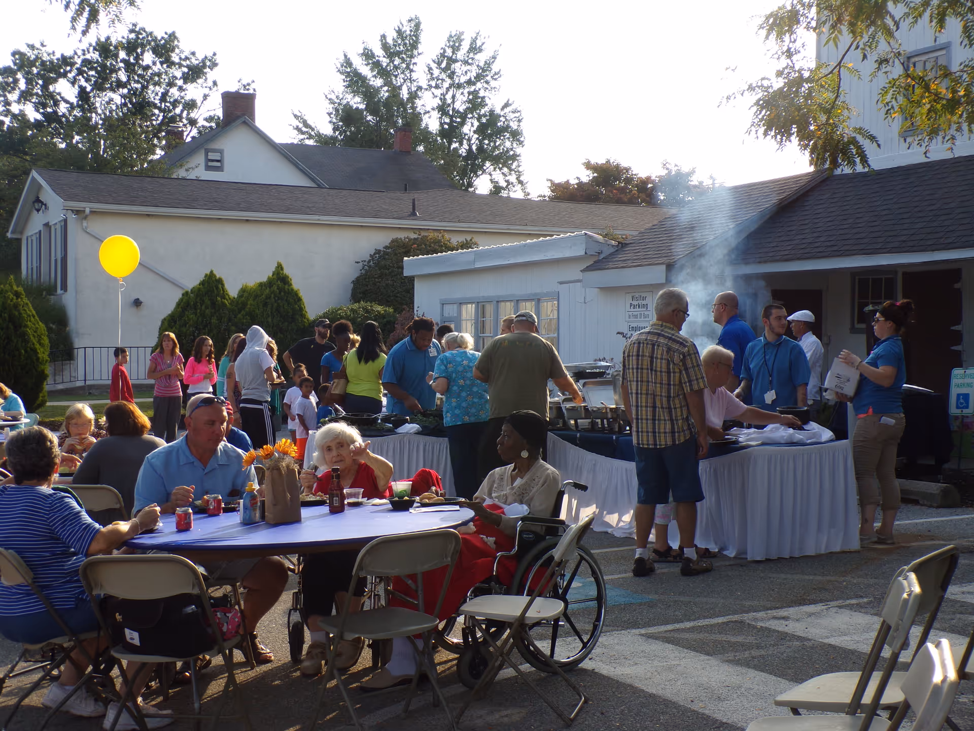 Outdoor gathering at Newport Meadows Nursing and Rehabilitation Center with people sitting at tables and others standing in line at a buffet table with food. There is a yellow balloon tied to a chair, and smoke is rising from the buffet area. The setting is outside near buildings with trees in the background.