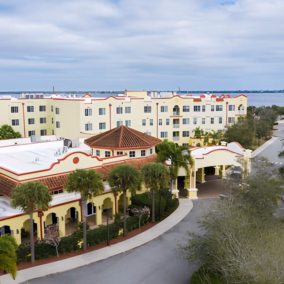 Aerial view of a Mediterranean-style multi-story senior living building with a covered porte-cochère, palm trees, and water in the background.