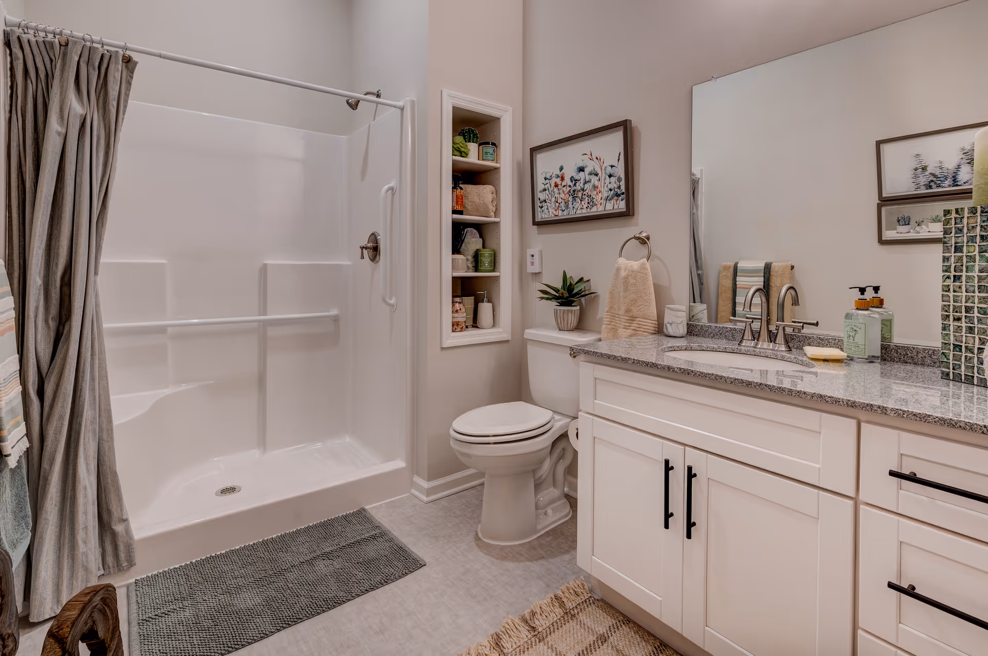 A clean and well-lit bathroom featuring a white shower with grab bars and a curtain, a toilet with a small plant and towel on the tank, a granite countertop with a sink, soap dispenser, and decorative items, and a large mirror above the sink. There are framed floral artworks on the walls and a built-in shelving niche with towels and toiletries.