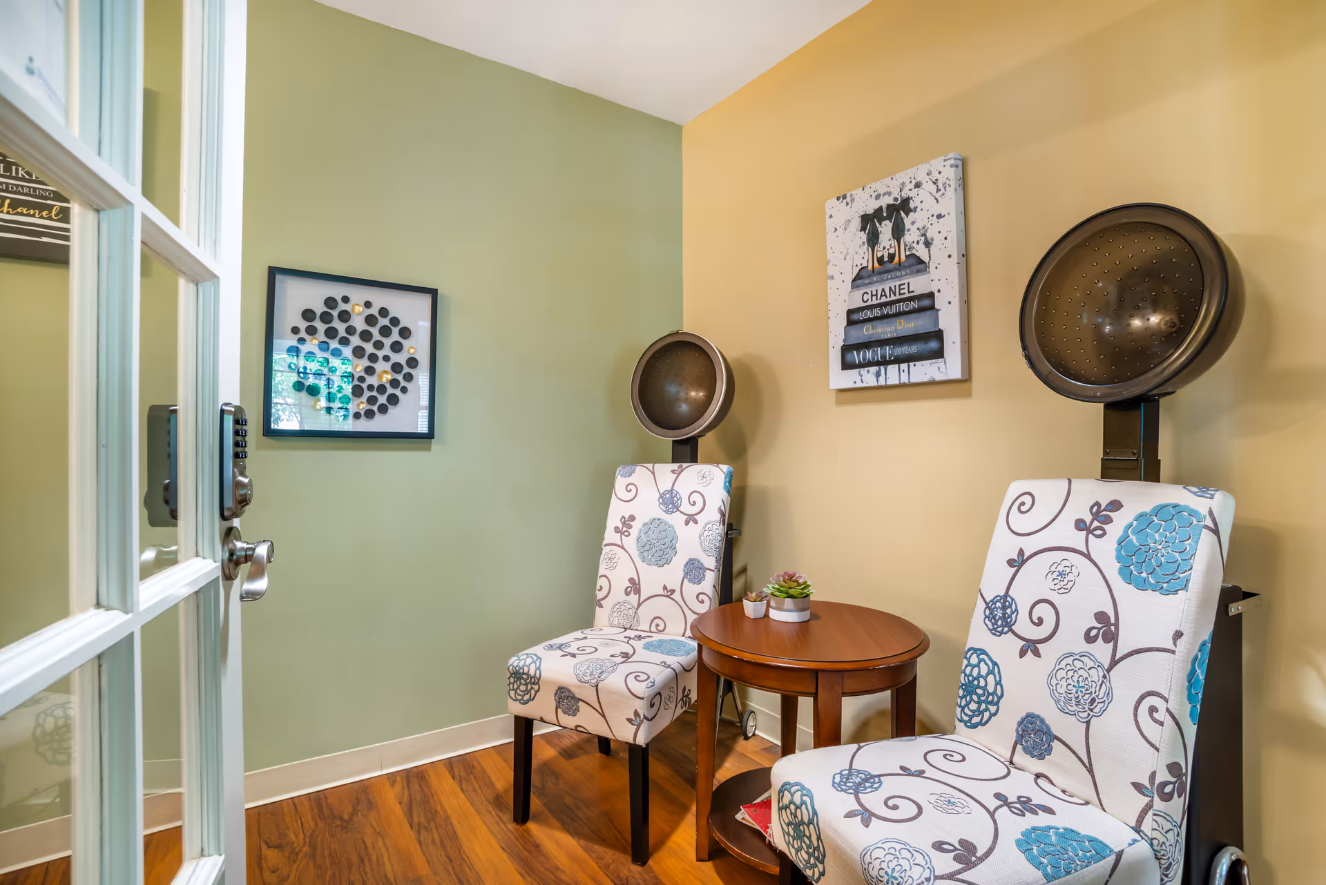 A cozy interior room with two floral-patterned chairs positioned around a small round wooden table with two small potted plants. Behind each chair is a vintage hair dryer hood. The walls are painted in two colors: light green on the left and beige on the right. Two framed artworks hang on the walls, one with a circular pattern and the other depicting stacked fashion books with a dog on top.