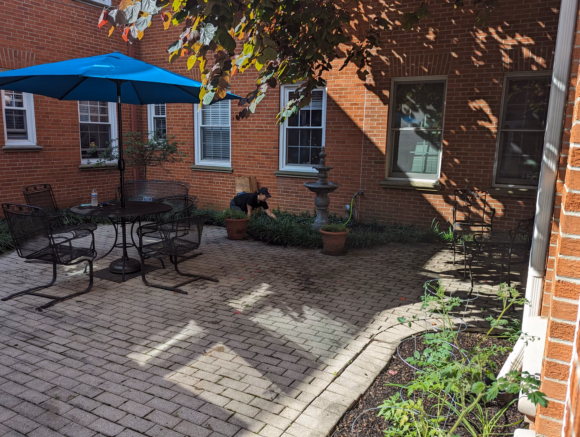 Sunny brick courtyard with a patio table and blue umbrella, potted plants, a fountain, and a person tending the garden.