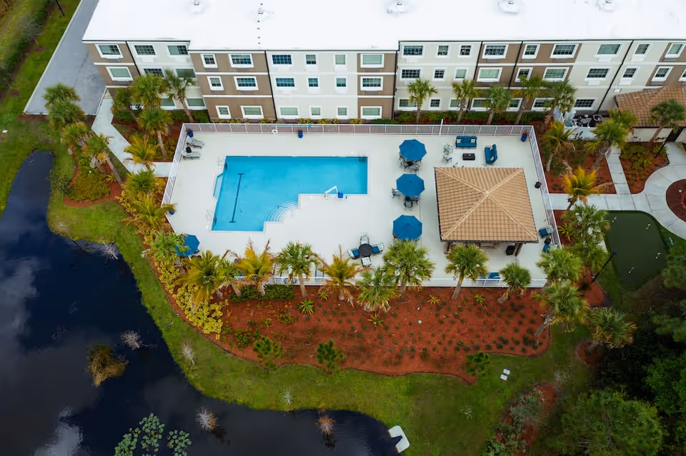 Aerial view of an outdoor swimming pool area at Discovery Village At Stuart, surrounded by a white fence. The pool area includes several blue umbrellas with tables and chairs, a covered pavilion, and landscaped greenery with palm trees. The pool is adjacent to a multi-story residential building, and there is a pond with lily pads nearby.