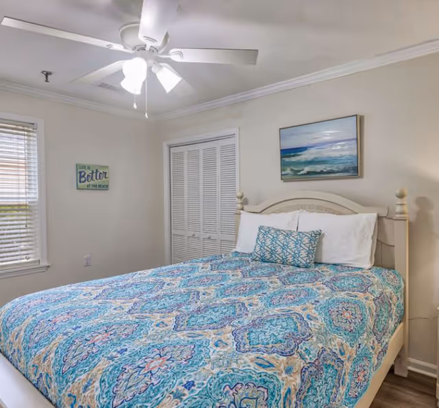 Sunlit bedroom with a bed dressed in a blue patterned quilt, ceiling fan, artwork, window blinds, and closet doors.